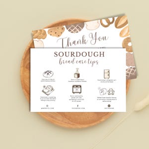 May include: A white card with "Thank You" and "SOURDOUGH bread care tips" in brown script. The card includes illustrated tips for bread care, such as storing and freezing. The card is on a wooden plate with bread illustrations.
