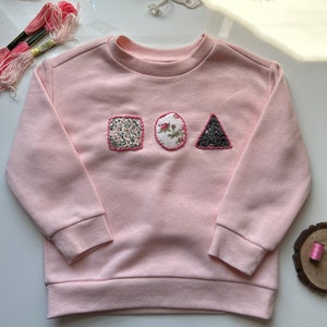 May include: A pink sweatshirt featuring a square, circle and triangle design crafted from fabric scraps. The shapes are adorned with floral patterns and sequins.