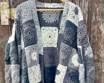 May include: A handmade, open-front crochet cardigan in a patchwork design. The sweater features a variety of gray and white squares, each with a floral motif. The cardigan is displayed on a wooden hanger against a weathered wooden background.