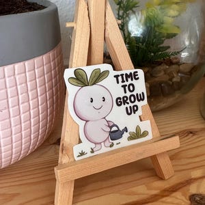 Peut inclure: Autocollant représentant un navet de dessin animé avec un arrosoir et le texte « TIME TO GROW UP ». L'autocollant est présenté sur un petit chevalet en bois. Un pot rose et gris et un terrarium en verre sont en arrière-plan.