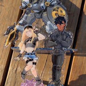 May include: A clear acrylic standee featuring a large grey mechanical robot with a red crest and gold shield, alongside two anime-style characters: one with long blonde hair in a grey top and blue shorts, and one with dark hair in a black suit.