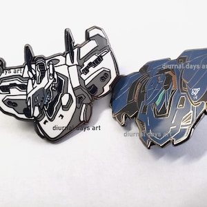 Puede incluir: Dos pines de esmalte que representan figuras robóticas. El primer pin es blanco con detalles grises y el segundo pin es azul con detalles grises. Ambos pines tienen un contorno negro.