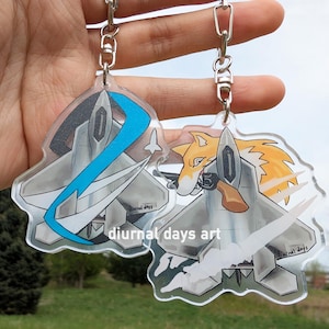 Op de afbeelding: Twee transparante acryl sleutelhangers met een cartoonwolf en een straaljager. De wolf is oranje en wit en houdt een pistool vast. De straaljager is grijs en heeft blauwe en witte accenten. De sleutelhangers zijn beide bevestigd aan zilveren kettingen.