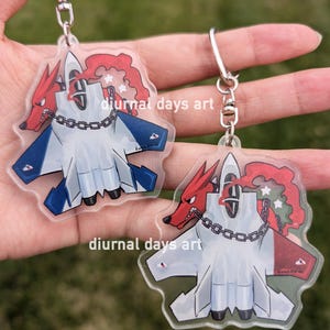 May include: Two clear acrylic keychains featuring a cartoon red fox with a chain around its neck, biting into a white fighter jet. The jet has red, white, and blue markings. The text "diurnal days art" is printed on the keychains.