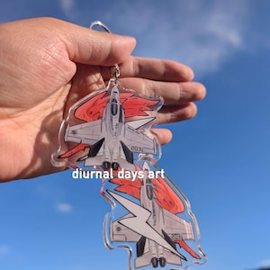 May include: A clear acrylic keychain featuring two gray fighter jet illustrations with red flame accents and the text diurnal days art. The jets are depicted from a top-down perspective with visible wings, engines, and the number 203.