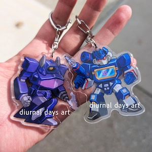 May include: Two clear acrylic keychains featuring cartoon robot characters. One is purple, the other is blue with a red and yellow screen. Both have silver key rings and the text "diurnal days art".