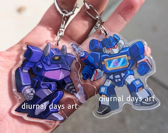 Può includere: Due portachiavi in acrilico trasparente con personaggi di robot dei cartoni animati. Uno è viola, l'altro è blu con uno schermo rosso e giallo. Entrambi hanno portachiavi argentati e la scritta "diurnal days art".