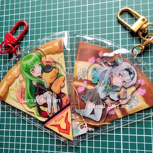 Może przedstawiać: Dwa przezroczyste breloczki do kluczy w kształcie kawałka pizzy z ilustracjami w stylu anime. Jeden przedstawia postać z zielonymi włosami, drugi z niebiesko-zielonymi włosami. Każdy breloczek ma inny kolor zapięcia. Widoczny jest tekst "diurnal days art".
