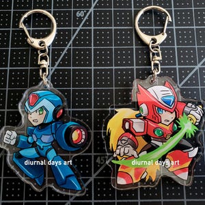 Puede incluir: Dos llaveros con personajes de anime de la serie Mega Man X. El llavero de la izquierda presenta a X, un robot azul con un casco rojo, en una pose de lucha. El llavero de la derecha presenta a Zero, un robot rojo con un casco blanco, sosteniendo una hoja de energía verde. Ambos llaveros tienen el texto "diurnal days art" en ellos.