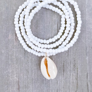 May include: White beaded necklace with a white cowrie shell pendant. The necklace is made of small, round beads and is layered for a full look.