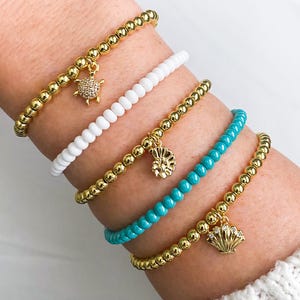 May include: A collection of beaded bracelets in gold, white, and turquoise. Each bracelet features a different charm: a turtle, a seashell, and a nautilus. The bracelets are worn on a wrist, showcasing their design and color.