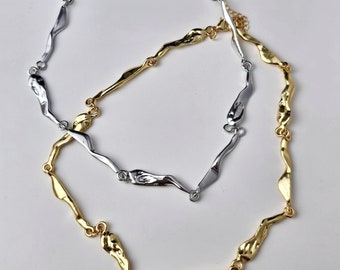 May include: A silver and gold chain necklace with a unique, abstract link design. The links are long and thin, and they are connected to each other in a way that creates a flowing, organic look.