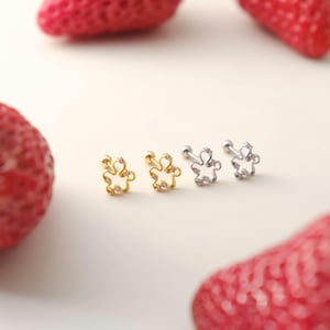 May include: Four flower-shaped stud earrings. Two are gold-toned with clear gemstones, and two are silver-toned with clear gemstones. The earrings are displayed on a white surface with red strawberries in the background.