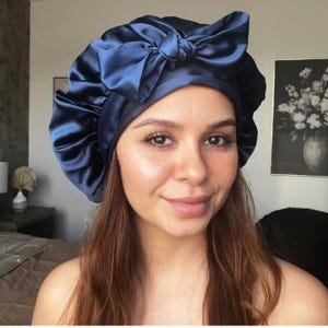 May include: A navy blue satin bonnet with a large bow on top. The bonnet is designed to protect hair while sleeping. The satin material has a slight sheen, and the bow adds a decorative element. The bonnet is a hair accessory.