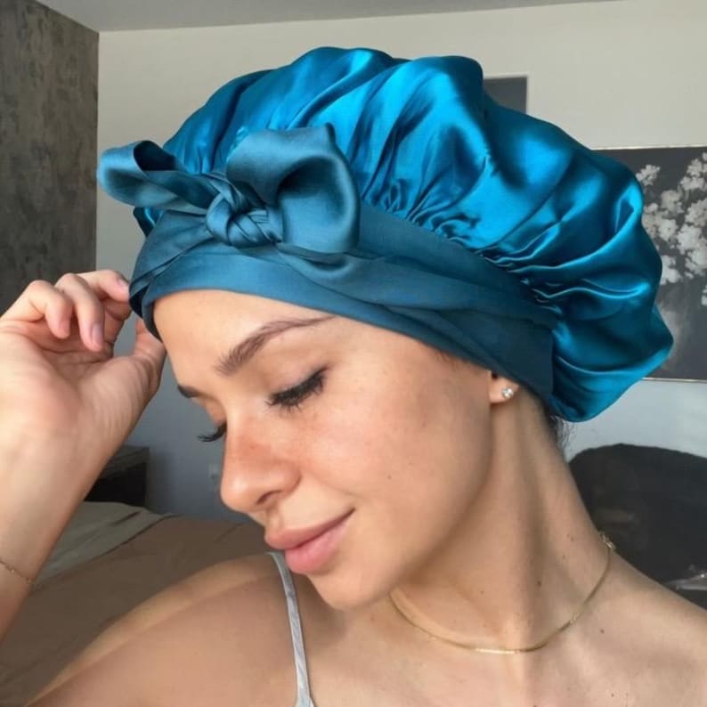 May include: A teal satin bonnet with a tied bow detail. The bonnet is designed to protect hair and maintain hairstyles. The shiny fabric reflects light, creating a smooth, elegant appearance. The bonnet is a hair accessory.