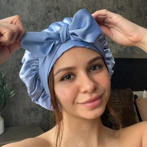 May include: A light blue satin bonnet with a large bow on top. The bonnet is designed to protect hair and is made of a shiny, smooth fabric. The bow is centered on the forehead.