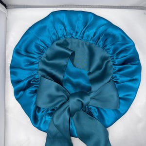 May include: A teal satin bonnet with a large bow. The bonnet is round with a gathered edge and a smooth, shiny texture. The bow is made of the same material and is centered on the front.