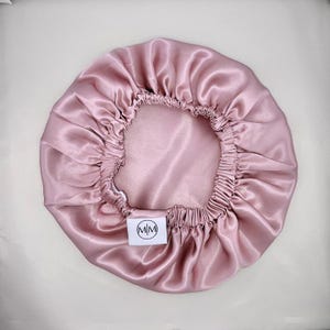 Peut inclure: Un bonnet en satin rose clair avec un élastique et une petite étiquette blanche avec les lettres "M/M" à l'intérieur d'un cercle. Le bonnet est conçu pour protéger les cheveux pendant le sommeil et est fait d'un tissu lisse et brillant.