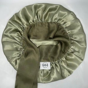 May include: Olive green satin bonnet with a matching tie. The interior is a darker olive green. A small white tag with the letters "MM" inside a circle is visible. The bonnet is designed to protect hair.