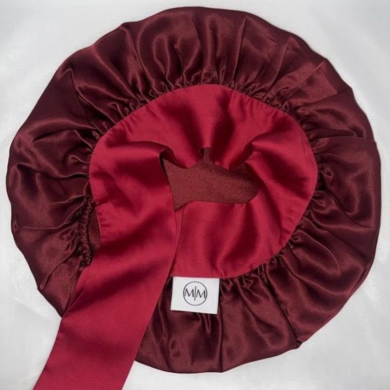 May include: A burgundy satin bonnet with a contrasting deep red interior and a long, matching tie. The bonnet features a gathered design and a small, white label with black text.