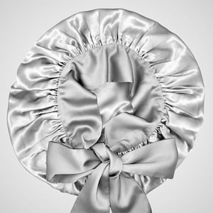 May include: A silver satin bonnet with a large bow at the front. The bonnet has a gathered elastic edge and a smooth, shiny texture. The bow is tied in a neat knot, adding a decorative element to the hair accessory.