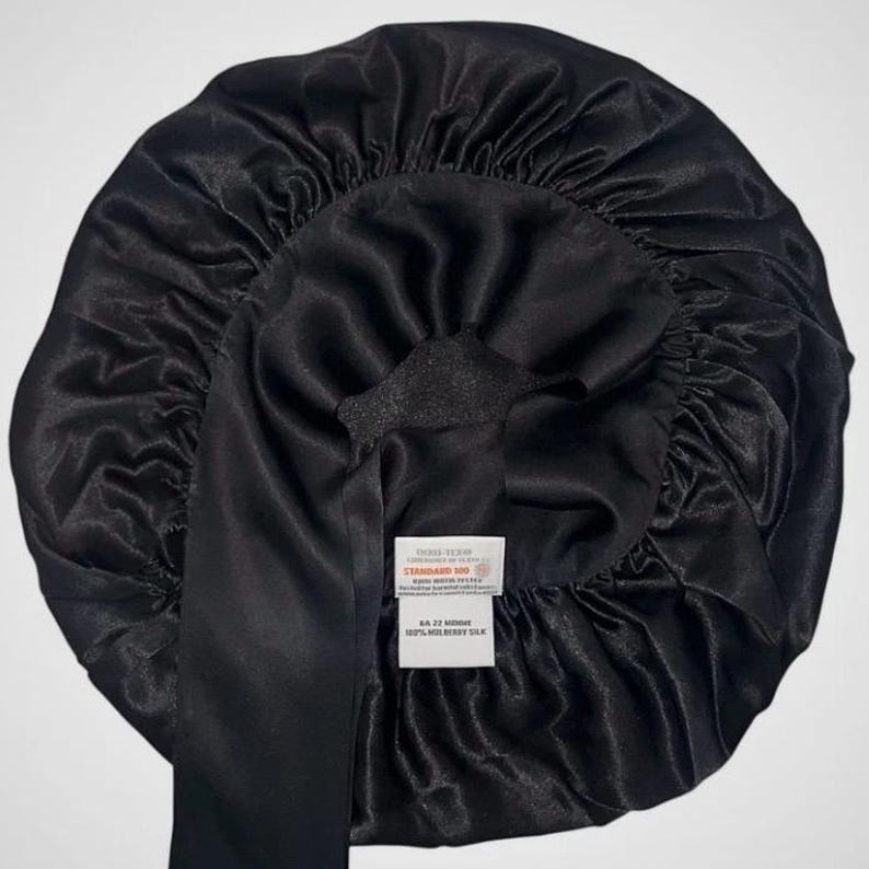 May include: Black silk bonnet with a satin finish. The bonnet has a wide band and a label that reads "STANDARD 100 by OEKO-TEX". The label also states "BA 22 MOME 100% MULBERRY SILK".