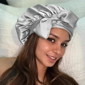 May include: A silver satin bonnet with a large bow. The bonnet is designed to protect hair while sleeping. The fabric has a slight sheen, and the bow is centered on the front. The bonnet is a light gray color.