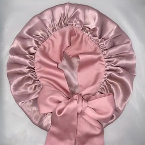 May include: A pink satin bonnet with a large bow. The bonnet is a light rose color on the outside and a darker pink on the inside. The bow is made of the same satin material and is tied in the front.