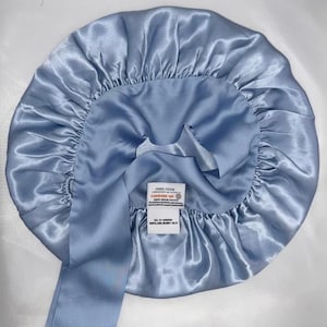 May include: A light blue silk bonnet with a long tie. The bonnet is round with a gathered edge and a label that reads "STANDARD 100" and "100% MULBERRY SILK". The fabric has a shiny, satin-like appearance.