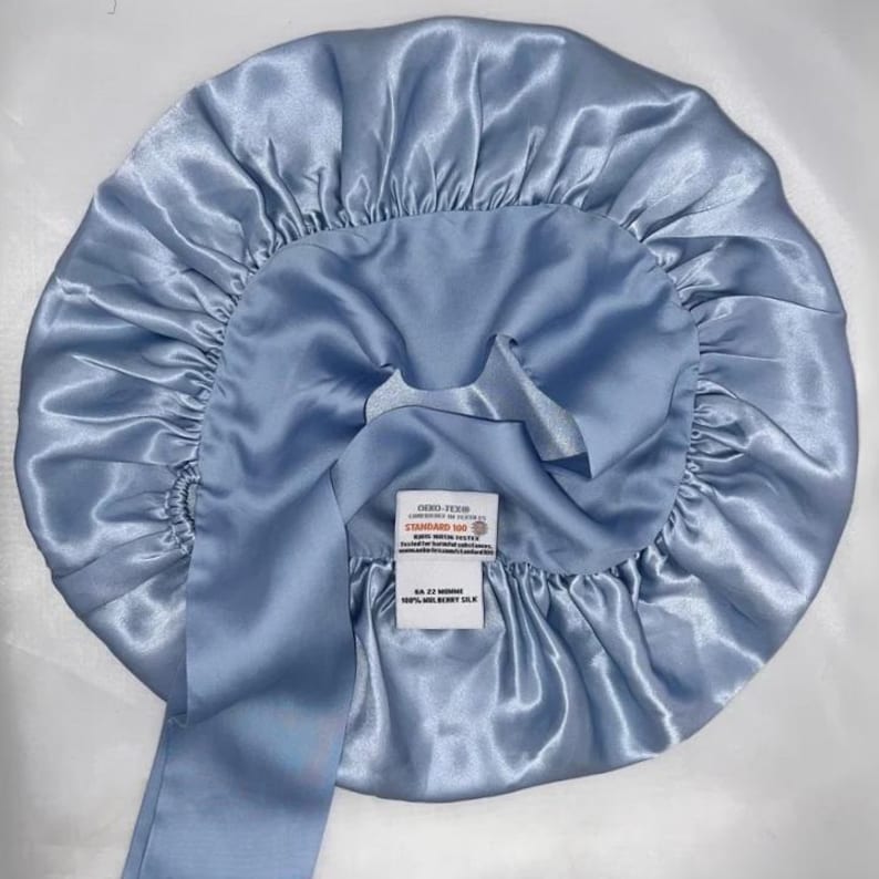 May include: A light blue silk bonnet with a long tie. The bonnet is round with a gathered edge and a label that reads "STANDARD 100" and "100% MULBERRY SILK". The fabric has a shiny, satin-like appearance.
