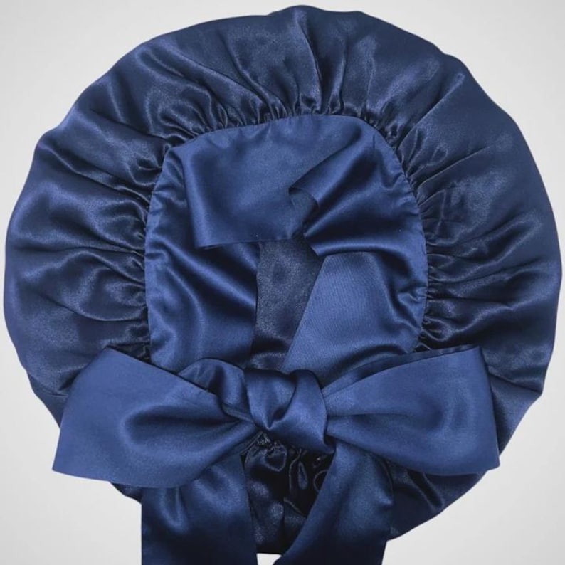 May include: A navy blue satin bonnet with a large bow at the front. The bonnet is gathered at the top and has a smooth, shiny texture. The bow is made of the same material and color as the bonnet.