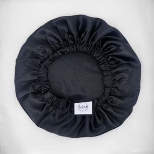 May include: A black satin bonnet with a gathered elastic edge. The bonnet is circular and features a small white tag with the letters "MM" inside a circle. The fabric has a shiny appearance.