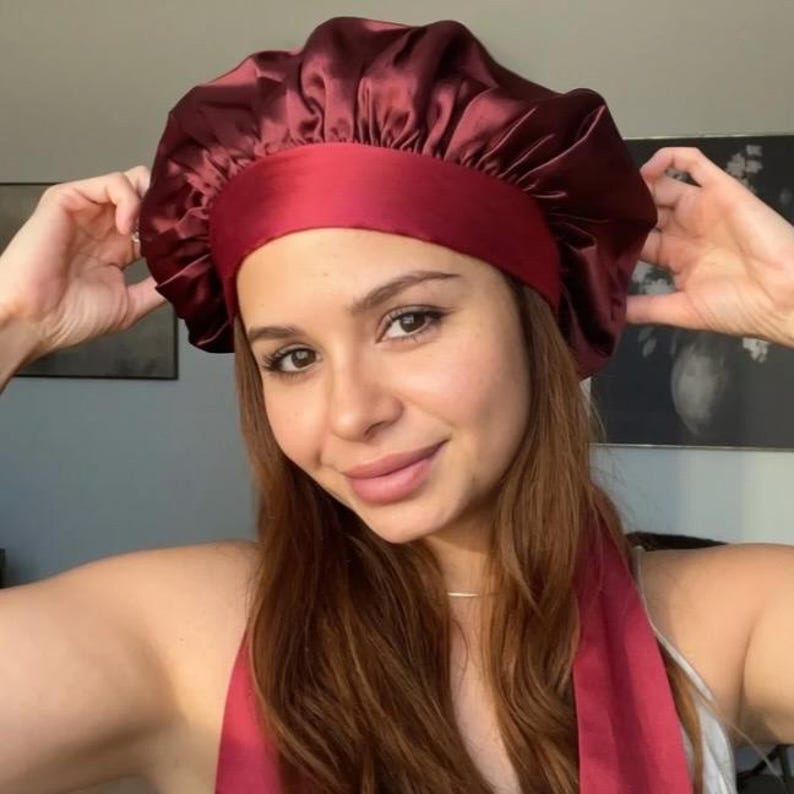 May include: Burgundy satin bonnet with a wide band and matching scarf. The bonnet is designed to protect hair while sleeping or during beauty routines. The fabric has a slight sheen, and the design is simple and elegant.