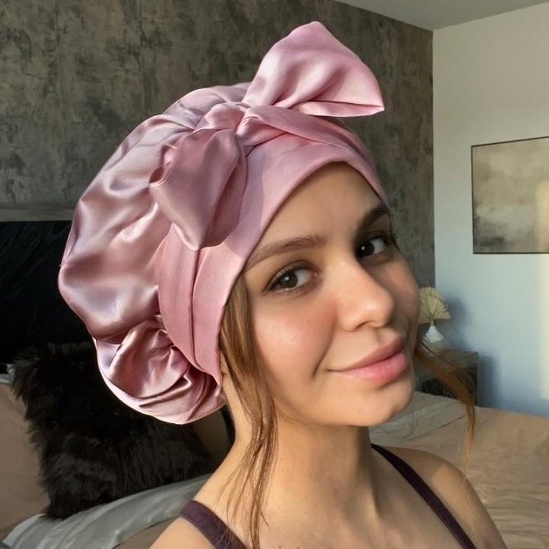May include: A pink satin bonnet with a large bow on top. The bonnet is designed to protect hair while sleeping. The satin material has a slight sheen, and the color is a soft, muted pink.