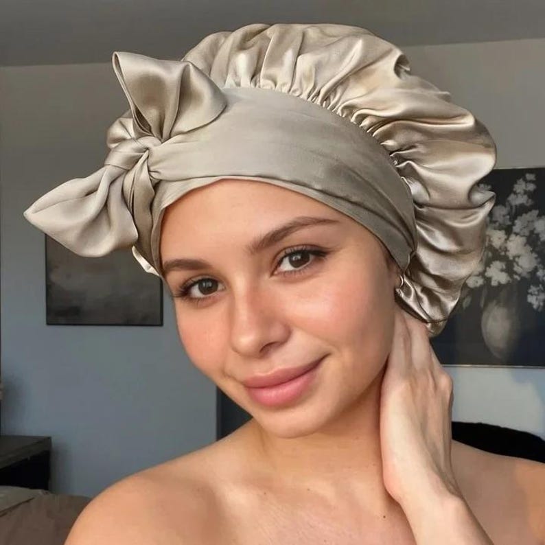 May include: A satin bonnet in a light champagne color. The bonnet has a gathered design and a large bow on the front. The material has a slight sheen, and the bonnet appears soft and smooth. The overall style is elegant and functional.