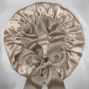 May include: A champagne-colored satin bonnet with a large bow. The bonnet is round with a gathered elastic edge. The bow is centered at the bottom of the bonnet and is made of the same satin material. The bonnet is designed to protect hair.