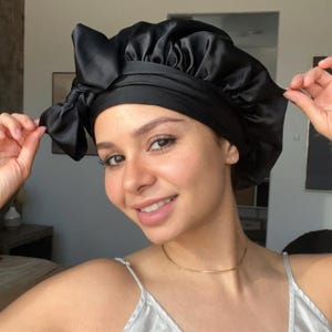 May include: A black satin bonnet with a large bow on the side. The bonnet is designed to protect hair and is worn on the head. The person in the image is smiling and wearing a light-colored camisole.