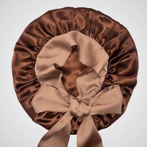 May include: A brown satin bonnet with a light brown interior and a large bow. The bonnet is designed to protect hair while sleeping. The satin material has a shiny, smooth texture.