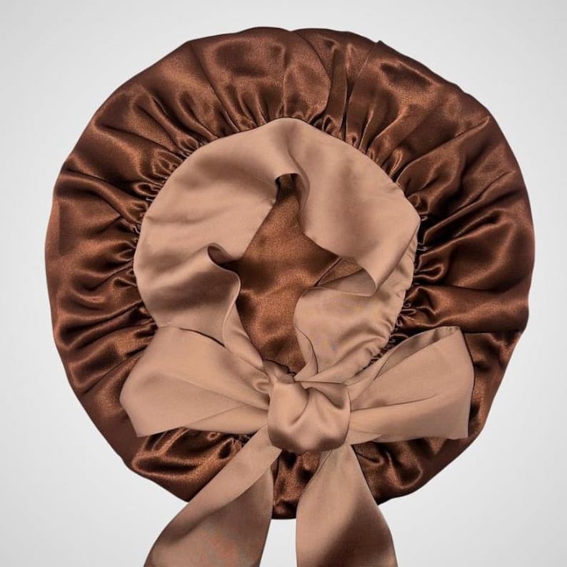 May include: A brown satin bonnet with a light brown interior and a large bow. The bonnet is designed to protect hair while sleeping. The satin material has a shiny, smooth texture.