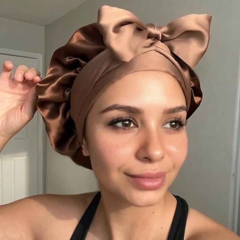 May include: A brown satin bonnet with a large bow on top. The bonnet is designed to protect hair and maintain hairstyles. The satin material provides a smooth surface to reduce friction and prevent hair breakage. The bonnet is a hair accessory.