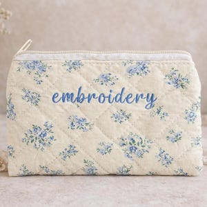 May include: Cream-colored quilted pouch with blue floral pattern and the word "embroidery" embroidered in blue script. The pouch has a zipper closure and is likely used for storing small items.