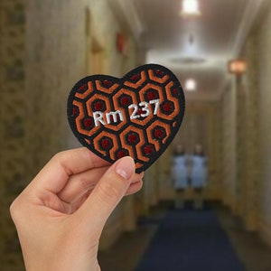 May include: A black heart-shaped patch with an orange and red geometric pattern. The patch has the text "Rm 237" embroidered in white.