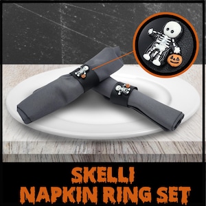 Può includere: Due tovaglioli di stoffa grigi con anelli per tovaglioli a forma di scheletro nero. Gli anelli presentano una piccola zucca e uno scheletro che tiene una zucca. Il testo "Skelli Napkin Ring Set" è stampato in arancione su una superficie di legno.