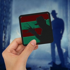 Op de afbeelding: Een geborduurde zwarte en groene patch met een silhouet van een persoon met een hoed en een mes. De patch heeft een rood-groen-zwarte gestreepte achtergrond.