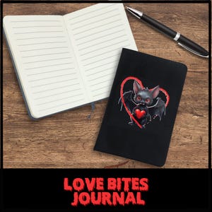 Puede incluir: Un diario negro con un lindo murciélago de dibujos animados que sostiene un corazón rojo. El diario tiene el texto "Love Bites Journal" impreso en la portada.