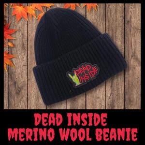 Könnte beinhalten: Schwarze Mütze aus Merinowolle mit umgeschlagenem Saum. Auf der Mütze befindet sich ein Aufnäher mit den roten Worten "DEAD INSIDE" und einer grünen Handgeste. Der Text "DEAD INSIDE MERINO WOOL BEANIE" ist unten rot dargestellt.