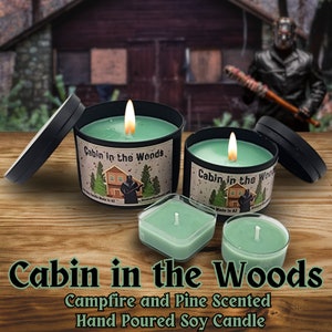 May include: A green soy candle with a black lid and a label that reads "Cabin in the Woods". The candle is hand-poured and scented with campfire and pine. The candle is sitting on a wooden surface.