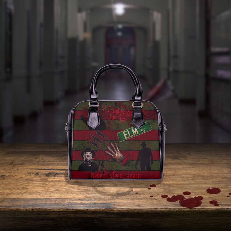 May include: A black handbag with a red and green striped design featuring Freddy Krueger from the movie A Nightmare on Elm Street. The bag has a black handle and a sign that reads "Elm St."