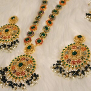 May include: A gold-toned jewelry set featuring a necklace and two pairs of earrings. The pieces are adorned with green, red, orange, and blue accents, and are embellished with black and white pearl-like beads. The jewelry is set against a white, fluffy background.