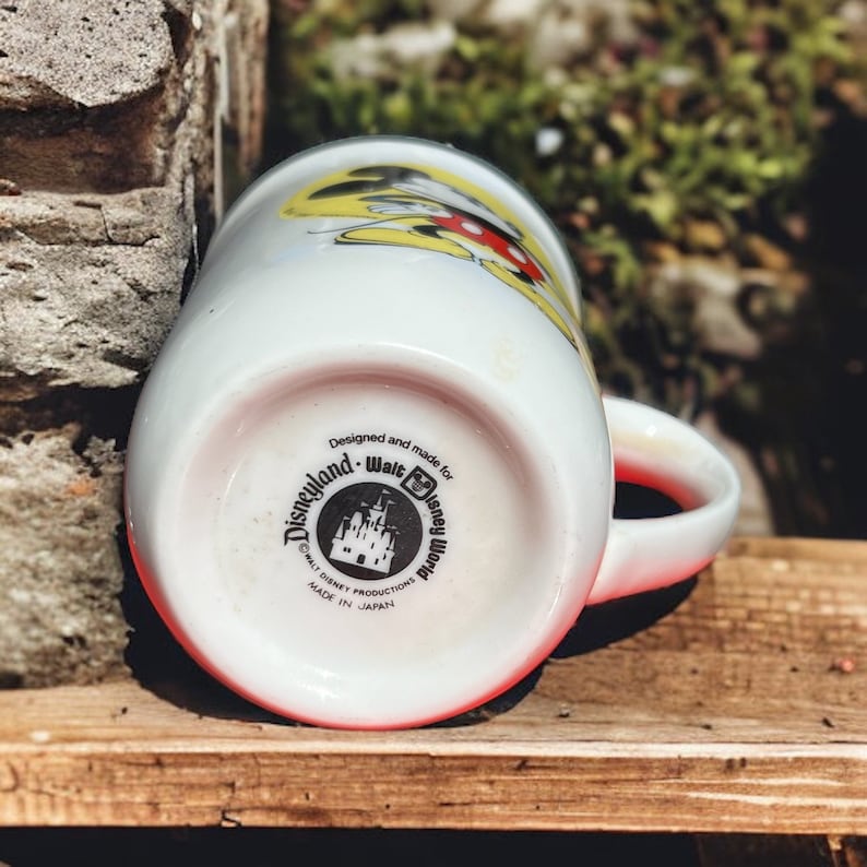 Peut inclure: Une tasse en c&eacute;ramique blanche avec un motif Mickey Mouse jaune, rouge et noir. Le fond de la tasse pr&eacute;sente un logo noir et blanc qui indique "Disneyland - Walt Disney World" avec un graphique de ch&acirc;teau et "&copy; WALT DISNEY PRODUCTIONS MADE IN JAPAN".